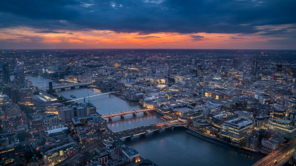 Aerial view of London at dusk with the Thames winding through the illuminated cityscape. St Paul's Cathedral is visible in the centre, with multiple bridges spanning the river. Dark blue skies contrast with a band of orange sunset and the glowing lights of buildings below.RetryClaude can make mistakes. Please double-check responses.