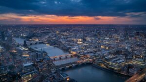Aerial view of London at dusk with the Thames winding through the illuminated cityscape. St Paul's Cathedral is visible in the centre, with multiple bridges spanning the river. Dark blue skies contrast with a band of orange sunset and the glowing lights of buildings below.RetryClaude can make mistakes. Please double-check responses.