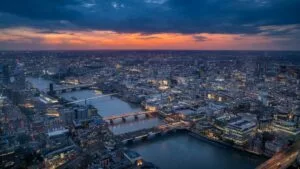 Aerial view of London at dusk with the Thames winding through the illuminated cityscape. St Paul's Cathedral is visible in the centre, with multiple bridges spanning the river. Dark blue skies contrast with a band of orange sunset and the glowing lights of buildings below.RetryClaude can make mistakes. Please double-check responses.
