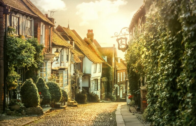 Charming cobblestone street lined with traditional British cottages featuring timber frames, thatched roofs, and ivy-covered walls in warm golden sunlight.