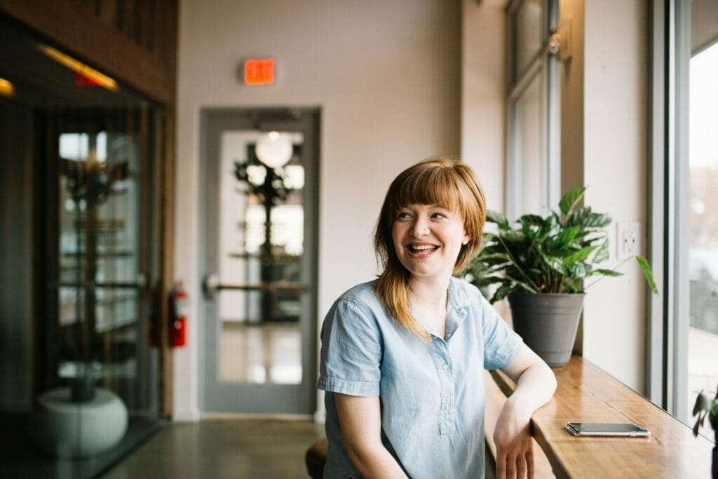 Professional woman in light blue shirt smiling whilst seated at wooden desk in modern office environment, representing HR professionals managing international assignments for their organisations.