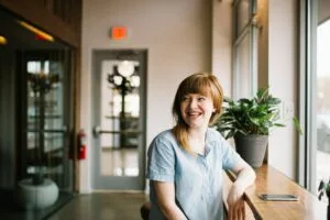 Professional woman in light blue shirt smiling whilst seated at wooden desk in modern office environment, representing HR professionals managing international assignments for their organisations.