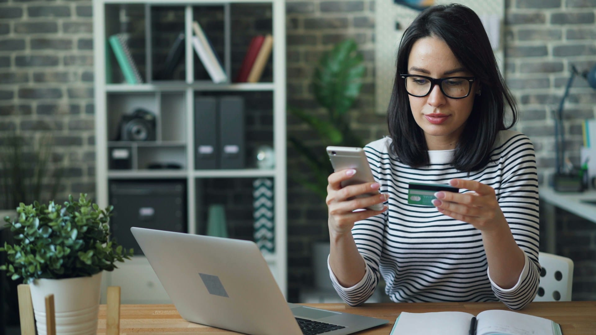 Professional woman in striped shirt using mobile phone and payment card at desk with laptop, illustrating digital expat financial services and online banking solutions.