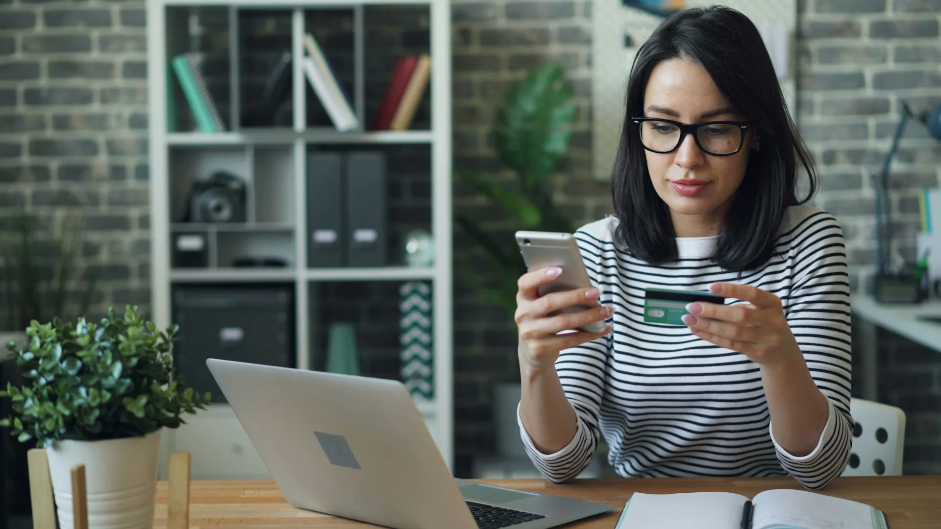 Professional woman in striped shirt using mobile phone and payment card at desk with laptop, illustrating digital expat financial services and online banking solutions.
